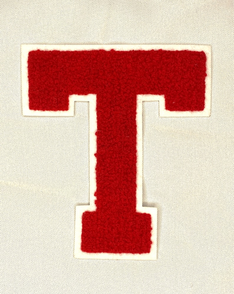 May include: A red felt letter "T" patch with a white border, set against a light gray fabric background. The letter has a textured, fuzzy appearance, typical of varsity letter patches. Ideal for jackets or bags.