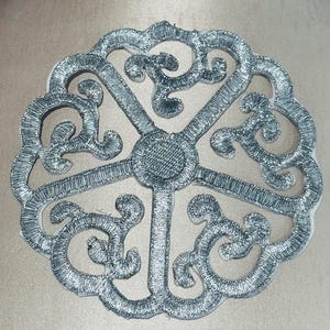May include: A silver embroidered applique with an ornate, symmetrical design. The circular shape features a central hub with radiating lines and swirling, decorative elements. The metallic thread creates a textured, shimmering effect.