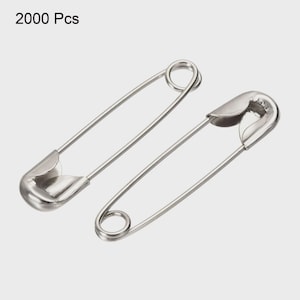 Pode incluir: Dois alfinetes de segurança prateados sobre um fundo claro. Os alfinetes são feitos de metal brilhante e têm um design clássico com uma extremidade enrolada e um fecho protetor. O texto "2000 Pcs" está no canto superior esquerdo.