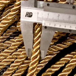 May include: A metallic gold twisted cord trim featuring a dense, spiraled texture. A silver metal caliper is positioned over a single strand of the cord, showing a measurement of approximately 0.25 inches in diameter for the individual rope.