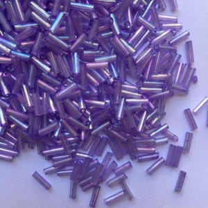Pode incluir: Um grande monte de contas de vidro roxo iridescentes, com algumas espalhadas sobre uma superfície branca. As contas são longas e finas, com uma forma ligeiramente arredondada.