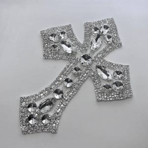 May include: A silver rhinestone cross applique with a sparkling design. The cross features a series of clear, faceted rhinestones in various shapes and sizes, set in a silver metal base. The cross is a decorative embellishment.