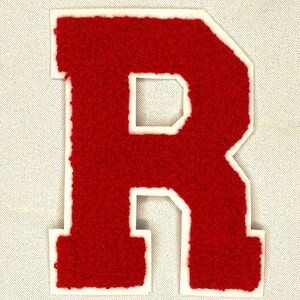 May include: A red, varsity-style letter "R" patch with a white border. The letter has a textured, fuzzy appearance and is set against a light-colored fabric background. This patch is suitable for jackets, bags, or other apparel.
