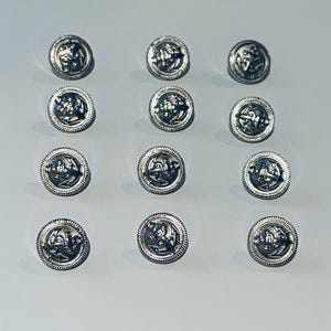 May include: Twelve silver-colored buttons with an anchor design. Each button has a rope-like border and a detailed anchor emblem in the center. The buttons are arranged in three rows of four on a light gray surface.