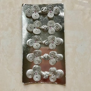 May include: Five silver Chinese knot buttons on a silver card. The buttons are made of braided cord and have a decorative knot in the center.