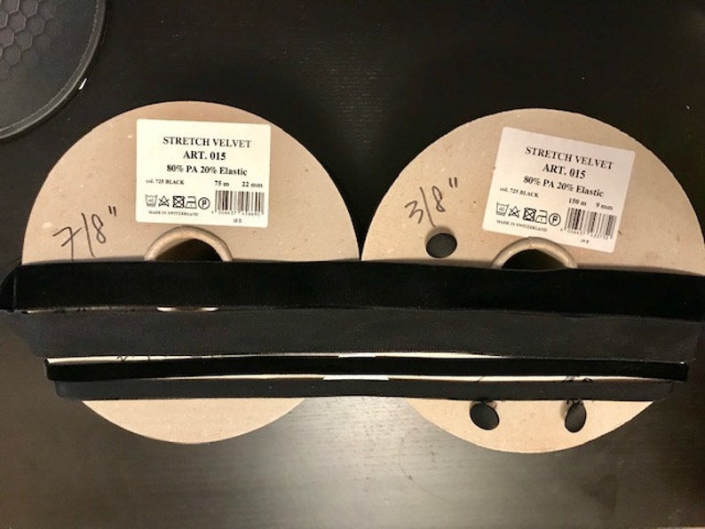 Pu&ograve; includere: Due bobine di tessuto in velluto stretch nero. Le bobine sono etichettate "Stretch Velvet Art. 015 80% PA 20% Elastico" e hanno le misure 7/8" e 3/8".
