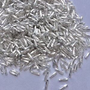 May include: A pile of silver glass beads, each bead is a small, thin cylinder.