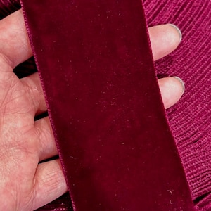 May include: A wide strip of deep burgundy velvet ribbon with a smooth, soft-touch pile surface. The ribbon features finished, stitched edges and is displayed against a background of matching burgundy pleated fabric with a vertical texture.