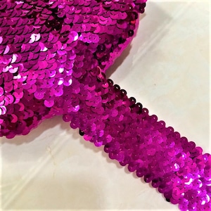 May include: A close-up of a pink sequined trim. The trim is made of small, round sequins that are sewn together in a continuous line.