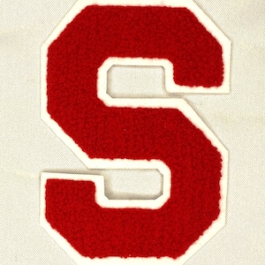 May include: A red felt letter "S" patch with a white border. The letter has a textured, fuzzy appearance and is sewn onto a light-colored fabric background. The patch is in a classic varsity style.