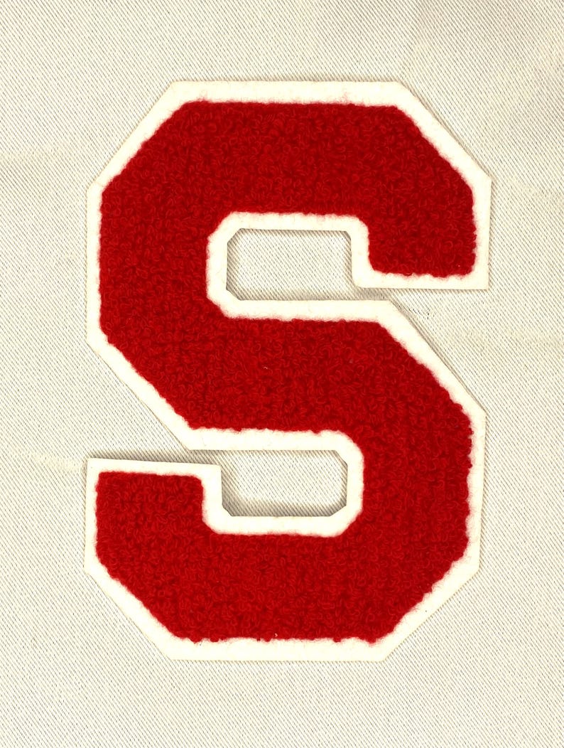 May include: A red felt letter "S" patch with a white border. The letter has a textured, fuzzy appearance and is sewn onto a light-colored fabric background. The patch is in a classic varsity style.