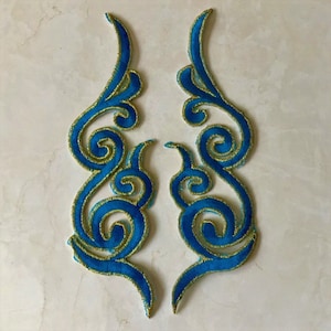 Puede incluir: Dos apliques simétricos y ornamentados con un diseño en espiral. Los apliques son de un azul vibrante con ribete dorado, con un patrón bordado detallado. El diseño recuerda a llamas estilizadas o adornos elegantes.