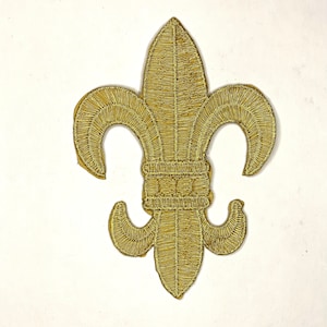 May include: Gold embroidered fleur-de-lis patch. The patch features a detailed design with a textured appearance. The emblem is a stylized lily, a symbol of French royalty, and is often used in heraldry and design.