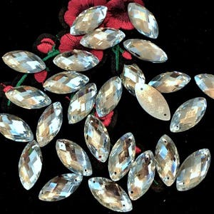 May include: Clear, faceted, marquise-shaped rhinestones scattered on a black background with red floral embroidery. Each rhinestone has a hole at each end. The rhinestones are reflective and catch the light.