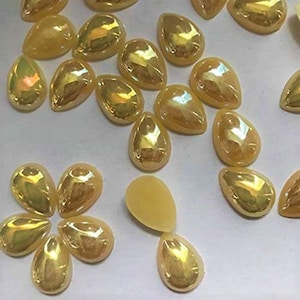 Pode incluir: Uma coleção de strass amarelos iridescentes em forma de lágrima. Os strass estão espalhados sobre uma superfície branca.