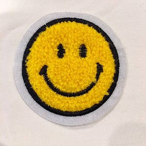 May include: Yellow smiley face patch with a black outline. The patch is made of fuzzy fabric and has a white border.
