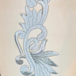 May include: A silver embroidered floral applique with a detailed, scrolling design. The applique features leaves and swirling patterns, perfect for embellishing garments or crafts. The embroidery is done in a metallic silver thread.