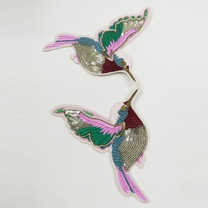 May include: Two embroidered hummingbird patches. The birds feature pink, green, blue, and gold sequins and beads. The patches have a white border and are set against a white background. The hummingbirds are in flight.