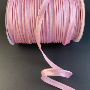 May include: A spool of pink satin ribbon with a stitched edge. The ribbon is wound around a cardboard spool with a white label.