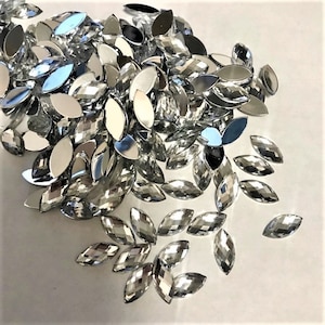 May include: A large pile of silver, faceted, marquise-shaped rhinestones. The rhinestones are scattered on a white surface.