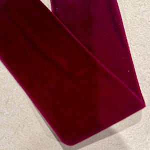 May include: A wide strip of deep magenta velvet ribbon with a soft, plush texture and finished edges. The fabric displays a rich, saturated color with subtle light reflections across its surface, showing a smooth, continuous length of material.
