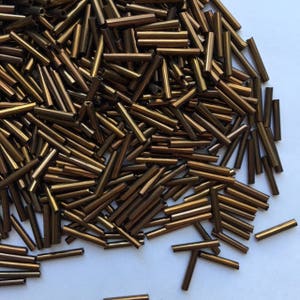 May include: A large pile of brown glass beads, all in the shape of long, thin cylinders. The beads have a metallic sheen and are scattered on a white surface.