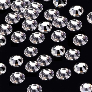 May include: A scattering of clear, round rhinestones on a black background. The rhinestones are various sizes and have a faceted, reflective surface.