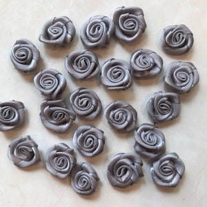 May include: A collection of 20 small, gray satin fabric roses. The roses are approximately 1 inch in diameter and have a delicate, ruffled appearance.