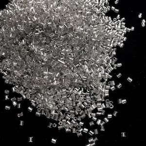May include: A pile of small, clear, silver-colored glass beads. The beads are cylindrical in shape and scattered across a black surface. These beads are suitable for jewelry making, crafts, and other creative projects.