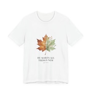 Può includere: Una t-shirt bianca a maniche corte con una grafica centrale di una foglia d'acero divisa tra toni arancioni autunnali e verdi, con una croce sovrapposta e piccoli rametti floreali. Sotto la foglia, il testo recita HE MAKES ALL THINGS NEW REV 21:5.