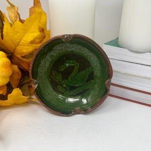 May include: A handmade ceramic ashtray with a green swan design in the center. The ashtray has a dark brown rim and a green interior. The ashtray is round and has a glossy finish. The ashtray is a decorative item.