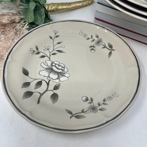May include: A round, beige ceramic plate featuring a dark gray floral illustration. The design includes a large central flower with leaves and smaller floral sprigs extending toward the edges. The plate has a scalloped rim with a thin dark gray line.