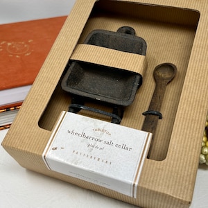 Puede incluir: Una caja de cartón marrón con una ventana transparente que muestra un salero de hierro fundido negro en forma de carretilla con una cuchara de madera. La caja tiene una etiqueta blanca que dice "Tabletop wheelbarrow salt cellar - pied de sel - Pottery Barn".