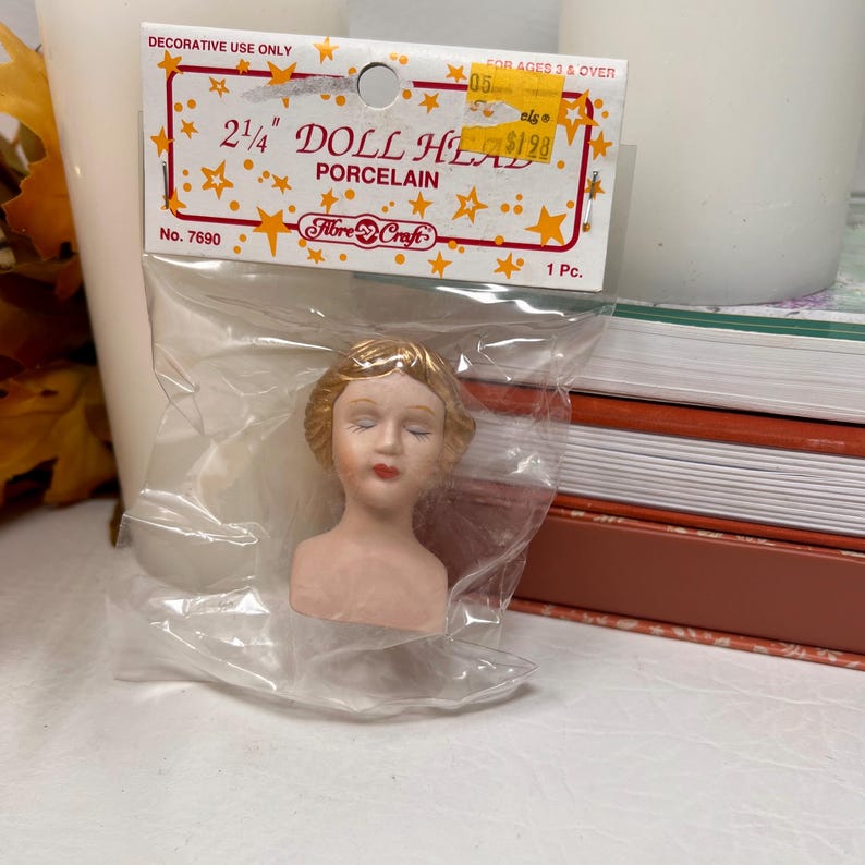 May include: A porcelain doll head, 2.25 inches tall, in a clear plastic bag. The doll head has closed eyes, red lips, and blonde hair. The packaging reads "DOLL HEAD PORCELAIN" and "Fibre Craft".