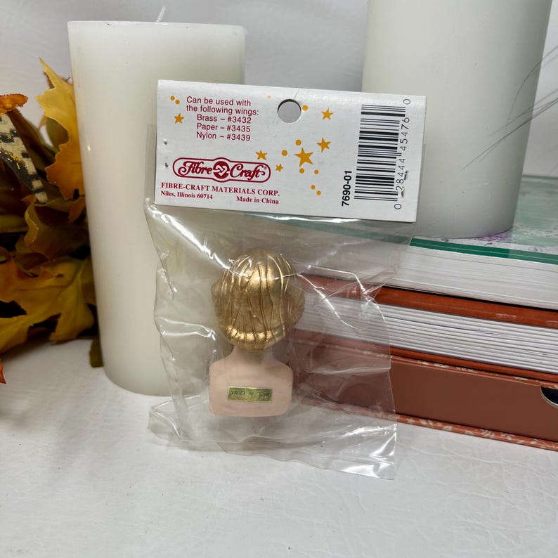 May include: A small, gold-colored bust figurine of a person with detailed hair, packaged in a clear plastic bag. The figurine is on a pink base. A product label is visible, with the text "Fibre-Craft Materials Corp."