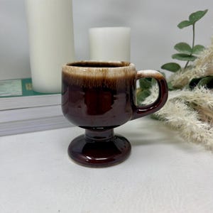 May include: A dark brown ceramic coffee mug with a pedestal base and a handle. The rim of the mug has a light brown glaze. The mug is on a white surface with candles and greenery in the background.