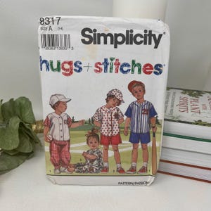 May include: A Simplicity sewing pattern envelope labeled 8317 Size A (1-4) Hugs + Stitches. The front features an illustration of three children wearing baseball-themed clothing, including a button-down shirt, shorts, and pants on a field.