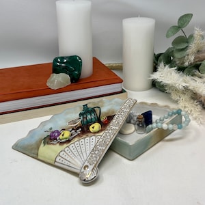 May include: A white porcelain trinket box with a hand-painted design of fruit and a fan. The box is open and reveals a blue and green interior with a small bottle and stones.