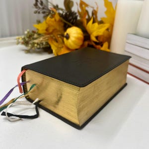 May include: A black book with gold-gilded edges and colorful ribbon bookmarks. The cover has a subtle embossed design. The book is placed on a white surface, with fall-themed decorations in the background.