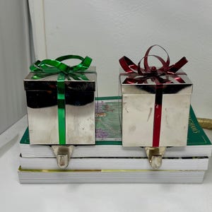 May include: Two silver Christmas stocking holders shaped like gift boxes, each with a ribbon bow. One has a green ribbon, the other a red ribbon. Both have a silver stocking hook. The boxes are sitting on a stack of books.