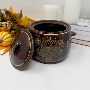 May include: A dark brown ceramic jar with a matching lid, featuring a gold-colored lattice pattern and small floral accents. The jar has a rounded body, a short handle on one side, and a gold rim detail on both the lid and the main container.
