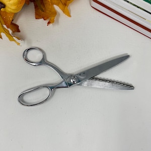 May include: Silver-colored, decorative scissors with a wavy blade edge. The scissors have a classic design with finger and thumb holes. The blades are open, showing the serrated edge. The scissors are likely used for crafting or fabric cutting.