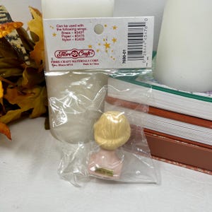 May include: A small doll head with blonde hair and a pink torso is packaged in a clear plastic bag. The packaging includes text from Fibre-Craft Materials Corp. and a barcode. The text says it can be used with brass, paper, and nylon wings.