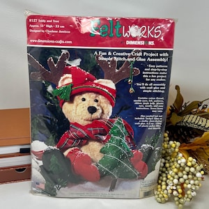 May include: Feltworks craft kit featuring a teddy bear wearing a red hat and scarf, sitting next to a green felt tree. The kit includes all the materials needed to create a 13-inch tall felt teddy bear and tree, including cotton thread, sparkle yarn, felt, patterns, ribbon, jingle bells, plastic eyes, a needle, and easy instructions.  The kit also requires Tacky Glue or a similar clear-drying craft glue, 6.5 oz. of plastic pellet filler, and fiberfill.