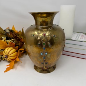 May include: A brass vase with a wide, flared rim and a rounded body. The surface is decorated with embossed fan-shaped patterns and inlaid with small, colourful stones in red, green, and blue, arranged in a vertical, cross-like design.