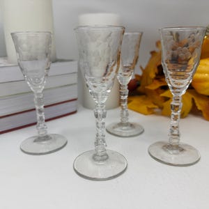 May include: Four clear glass cordial glasses with etched floral patterns on the bowls. Each glass features a slender stem with a faceted, multi-tiered design and a circular base. The glass material is transparent and smooth throughout the structure.