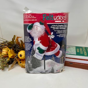May include: A felt craft kit to make a Santa Claus figurine. The kit includes felt pieces, embroidery floss, and instructions. The Santa is 18 inches tall and is holding a list. The kit is packaged in a clear plastic bag with a colorful label that says "Feltworks by Dimensions".