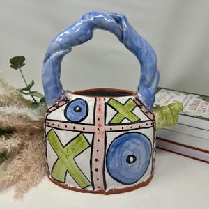 May include: A ceramic teapot featuring a white base with a hand-painted tic-tac-toe grid in pink. The design includes large blue circles and green X marks. It has a twisted blue handle and a small green spout extending from the side of the body.
