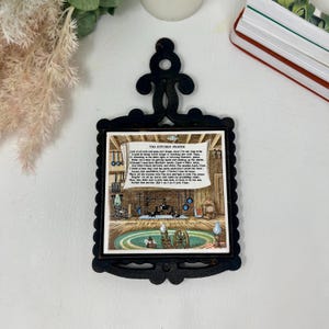 May include: Black cast iron trivet with a decorative top and a framed ceramic tile. The tile features a vintage illustration of a kitchen scene with the text "THE KITCHEN PRAYER". The trivet is designed to protect surfaces from hot cookware.