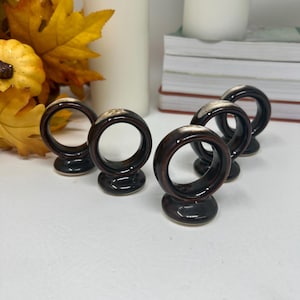 Può includere: Set di cinque anelli portatovaglioli in ceramica. Ogni anello è marrone scuro con un accento più chiaro, bianco sporco sulla parte superiore. Gli anelli sono circolari e poggiano su una piccola base rotonda. Lo sfondo include foglie autunnali e candele.